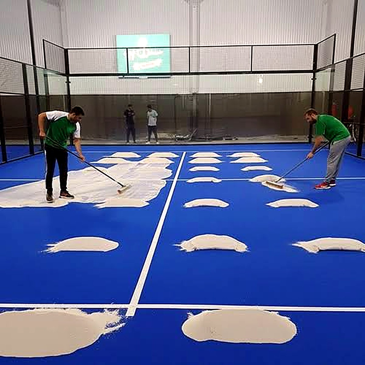 Két szakember keféli be a töltőanyagot a kék padel műfűbe a frissen fektetett pályán