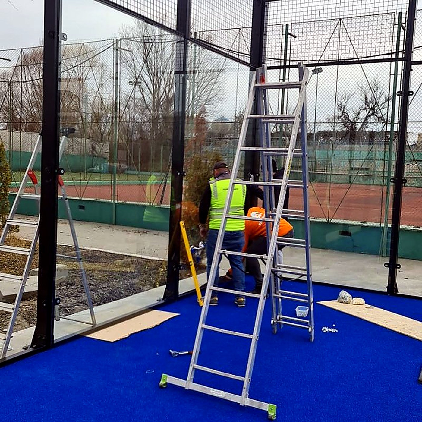 Kültéri padel pálya üvegfalának szerelése létrákról: szerelők láthatósági mellényben rögzítik az edzett üvegpanelt